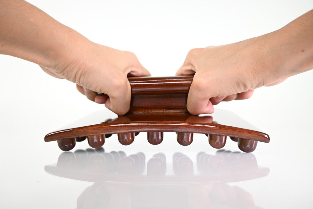 Two hands holding a dark brown Mushroom Massager with 48 fingertip-like nodes for cellulite massage and body sculpting tools. Made of pine wood in Colombia, 9 inches long and 3 inches tall.